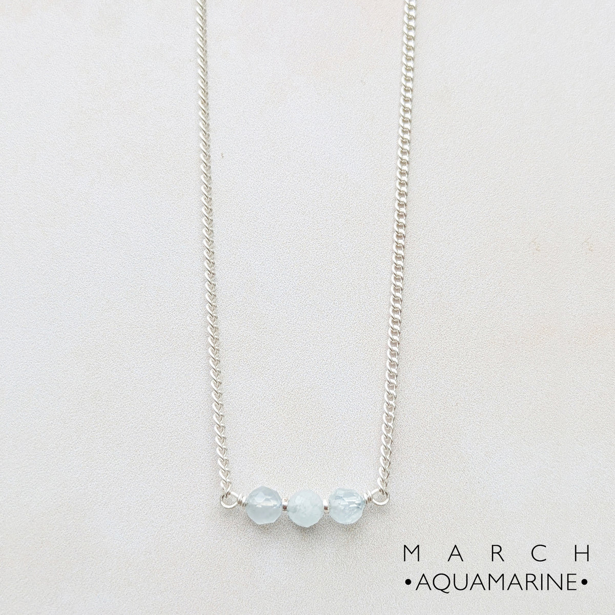 A silver chain birthstone necklace featuring three small, faceted light blue aquamarine beads on a delicate silver bar, displayed against a clean white background with 'MARCH • AQUAMARINE •' text in the lower right.