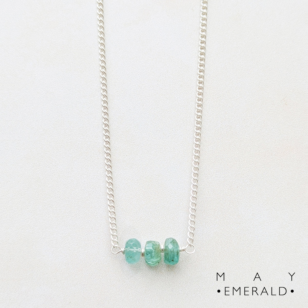 A silver birthstone bar necklace featuring three small, faceted Emerald gemstone beads at the centre, displayed against a clean white background with the brand name 'MAY EMERALD' in the bottom right corner.