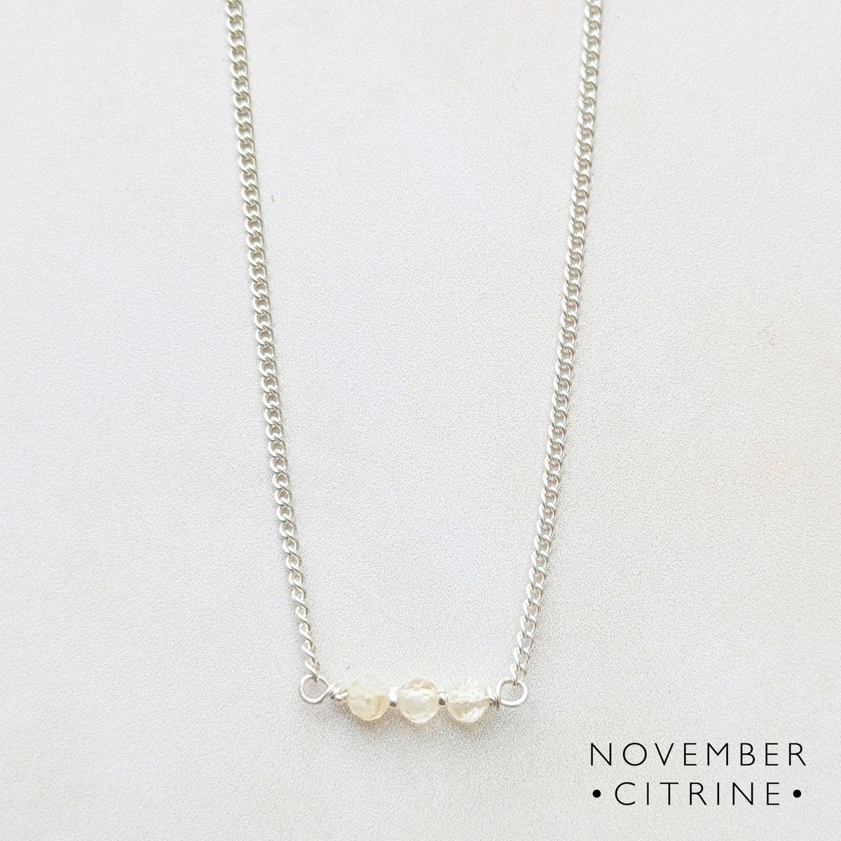 A delicate silver birthstone bar necklace featuring three small citrine gemstones at the centre, displayed on a light off-white background with the text  'NOVEMBER CITRINE' printed in the bottom right corner.
