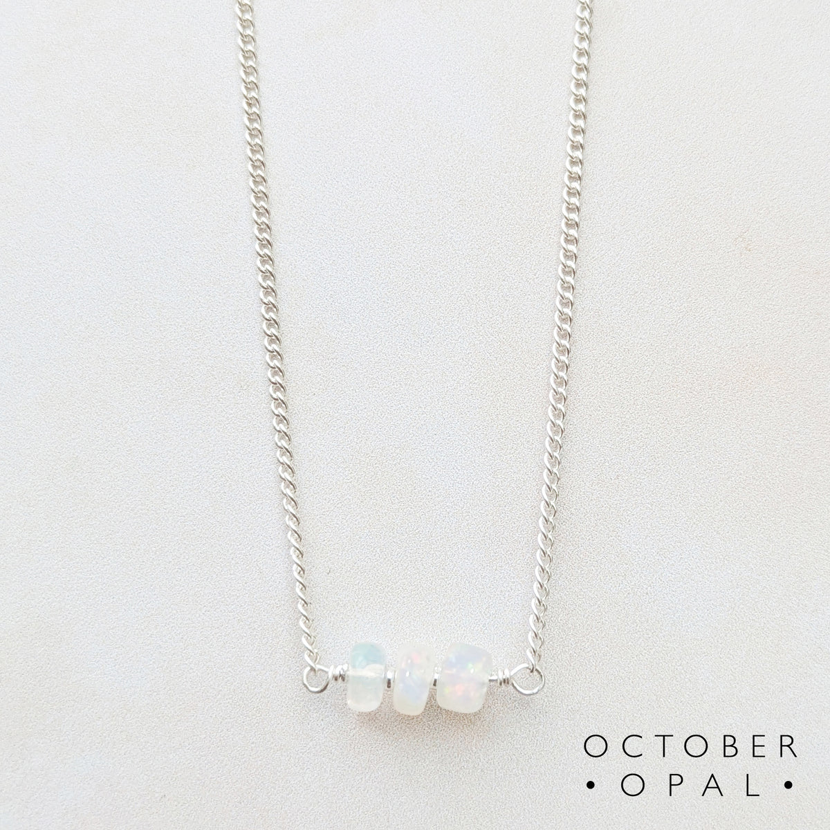 A delicate silver birthstone bar necklace featuring three small, round, opalescent beads in the center, displayed on a clean white background with the text 'OCTOBER OPAL' printed in the bottom right corner.