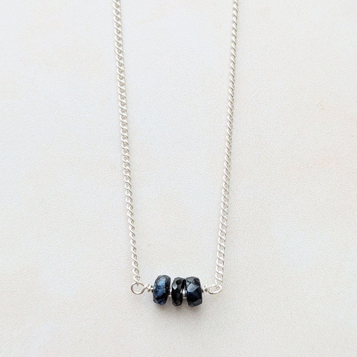 A delicate silver chain necklace featuring three small, dark blue faceted Sapphire beads clustered at the centre, displayed on a clean, light off-white background.