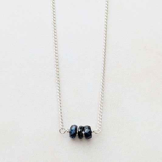 A delicate silver chain necklace featuring three small, dark blue faceted Sapphire beads clustered at the centre, displayed on a clean, light off-white background.