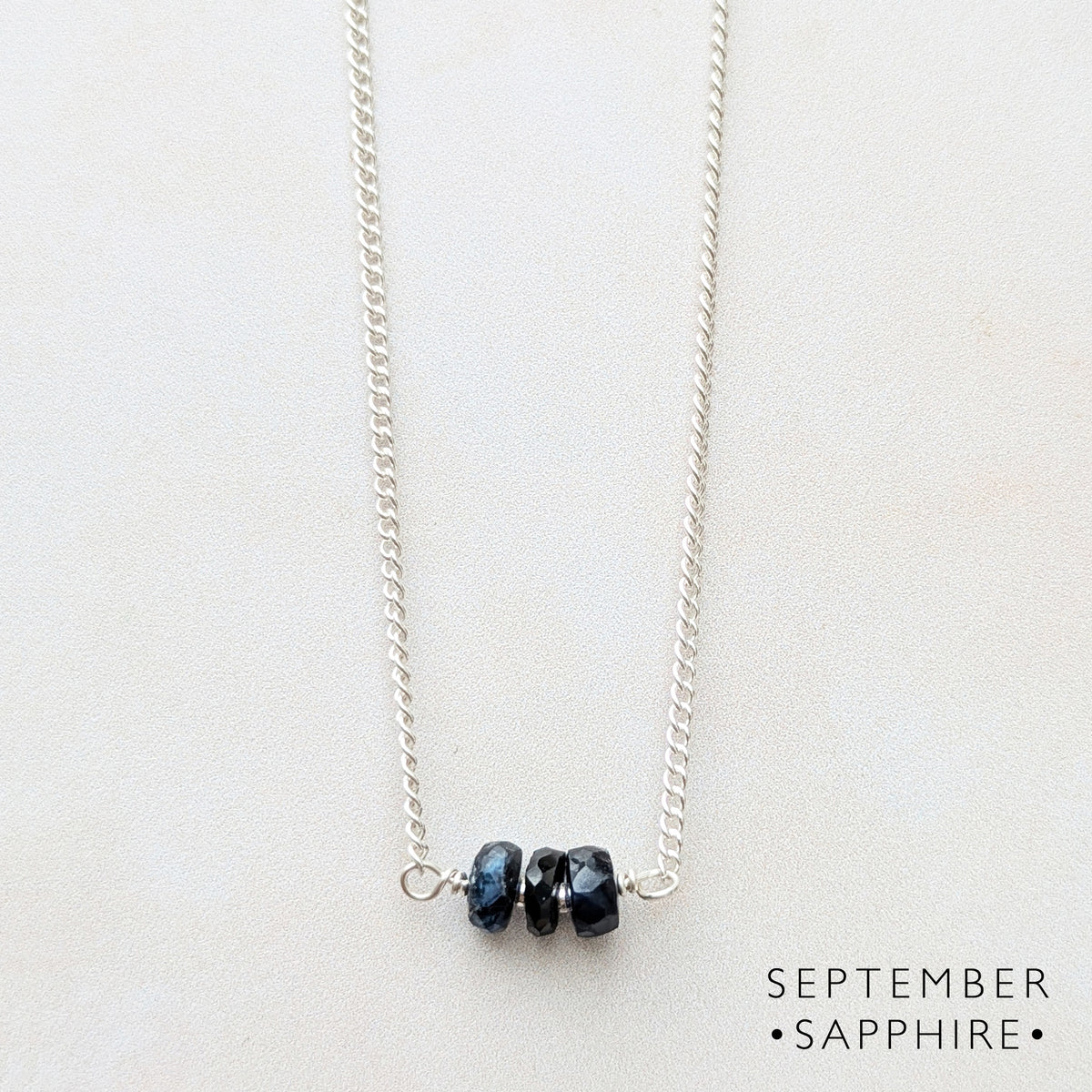 A silver birthstone bar necklace featuring three small, faceted dark blue gemstone beads on a light cream background, with 'SEPTEMBER SAPPHIRE' text in the bottom right corner.