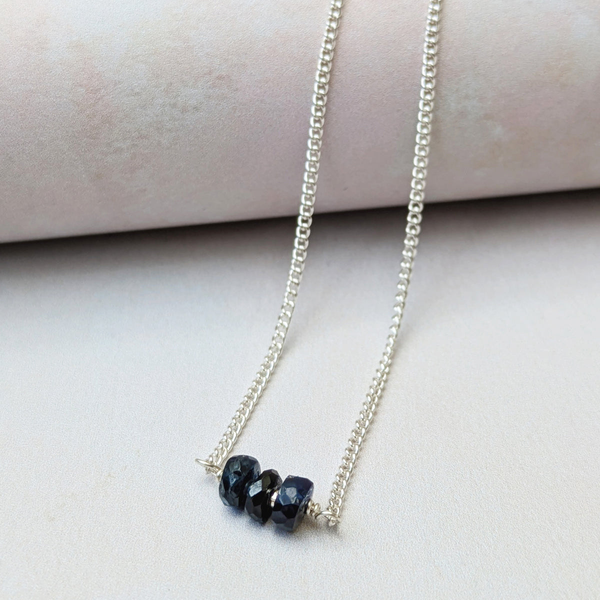 A silver chain September birthstone necklace with three small, faceted dark blue Sapphire beads, displayed on a light pink cylindrical jewelry stand against a soft beige background.