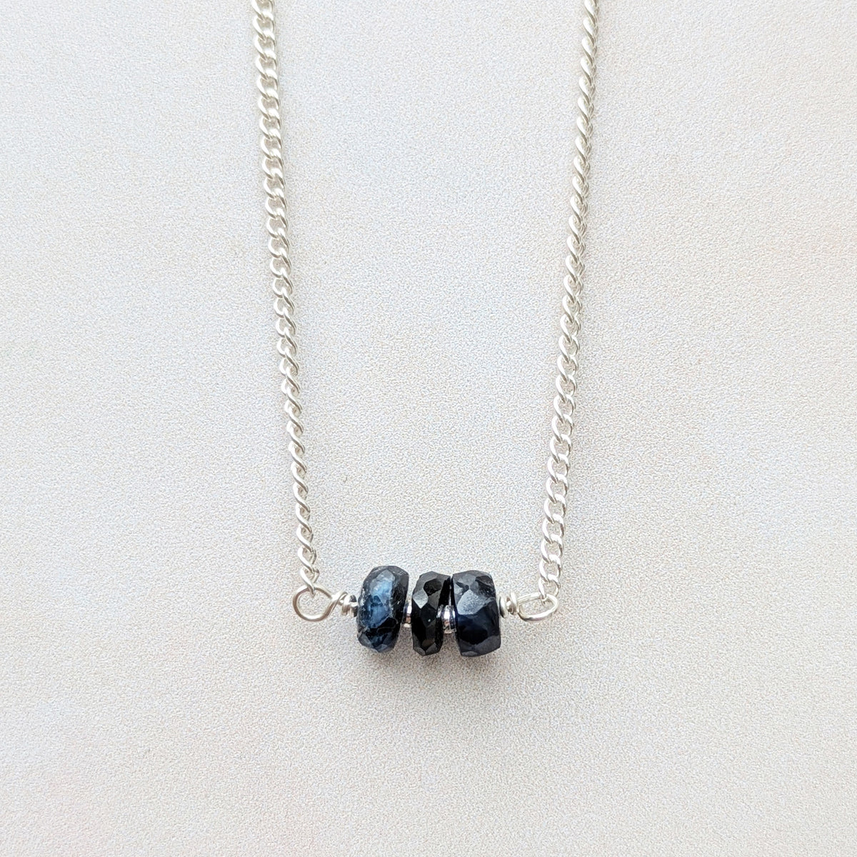 A silver birthstone necklace for September,  featuring three small, faceted dark blue Sapphire gemstone beads at the centre, displayed on a light gray background.