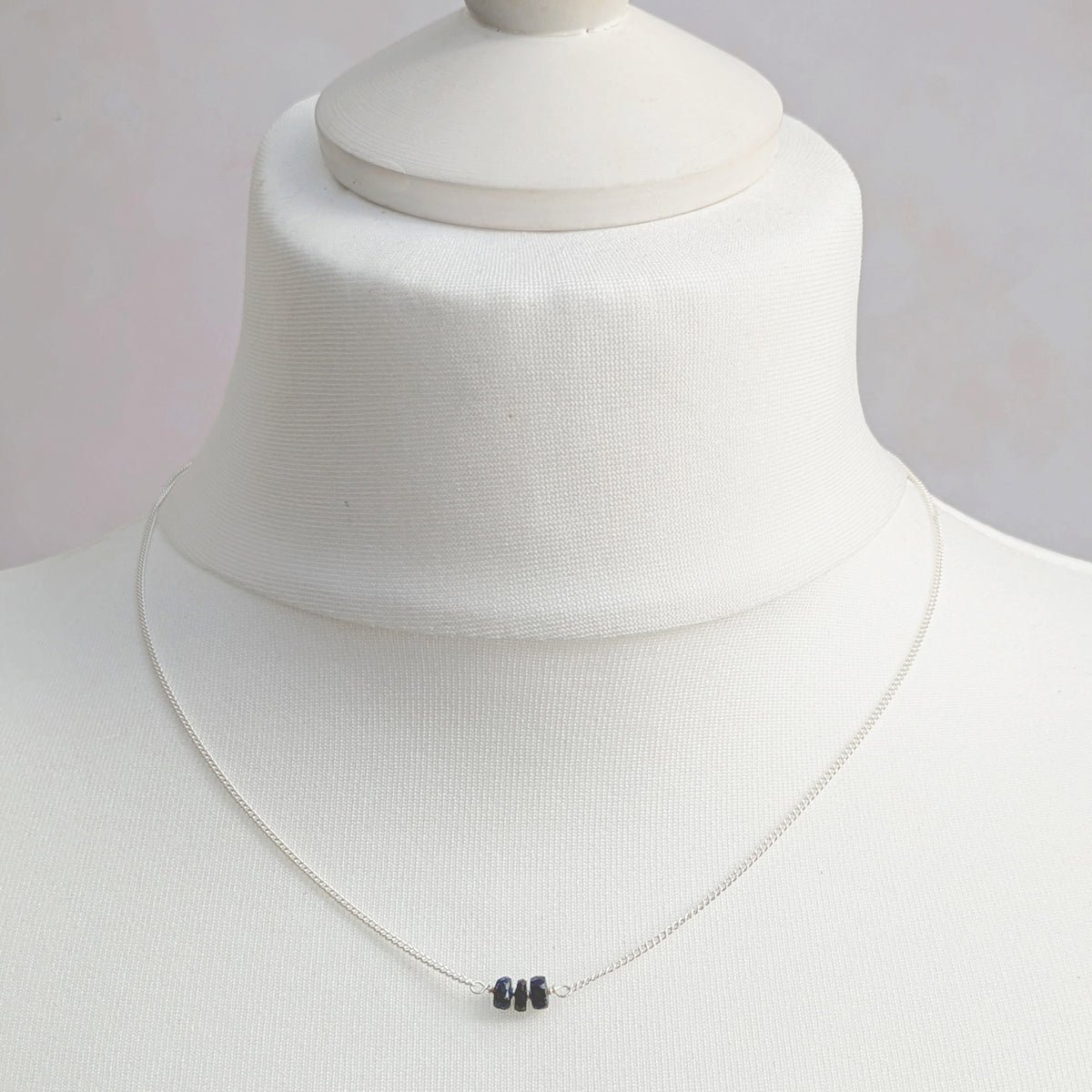 A delicate silver chain necklace with three small dark blue Sapphire beads at the centre, displayed on a white fabric mannequin bust.