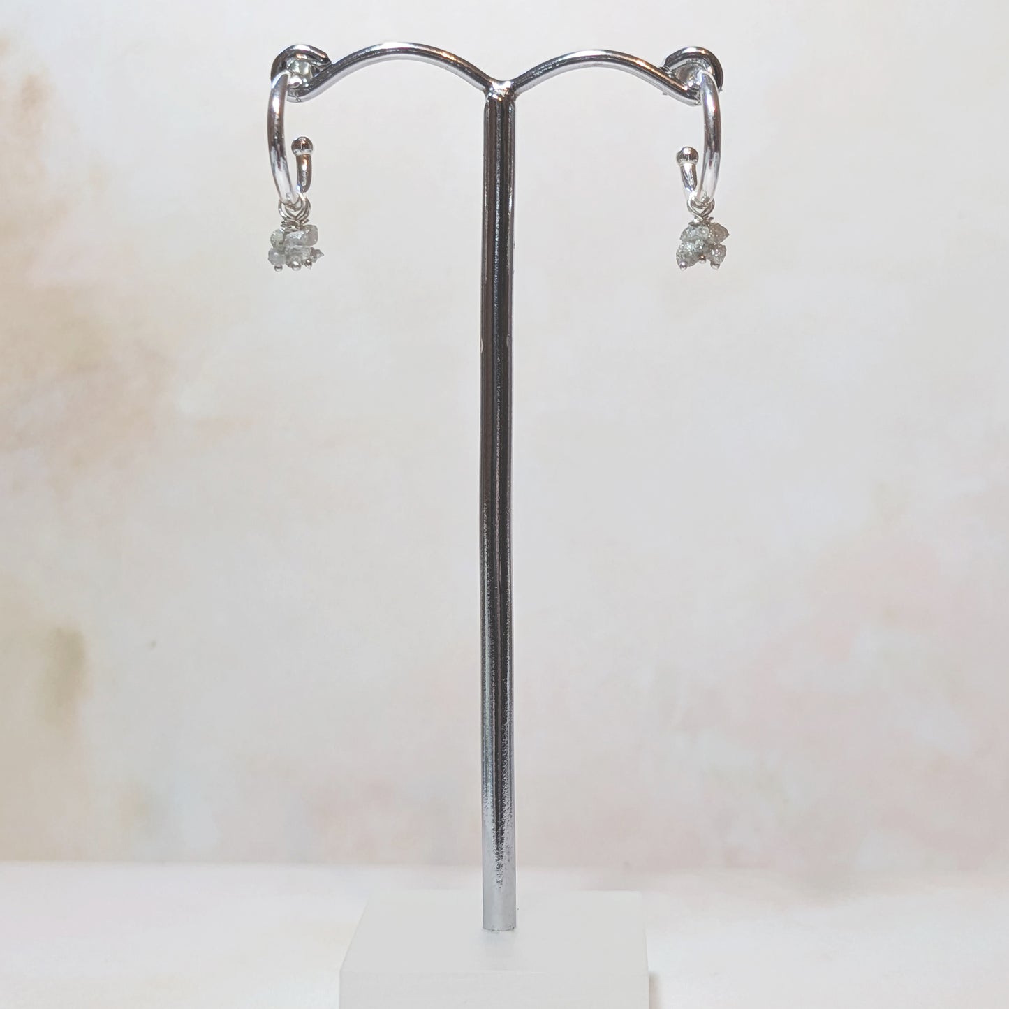 Silver hoop earrings with rough Diamon nugget charms on a chrome stand against a white background.