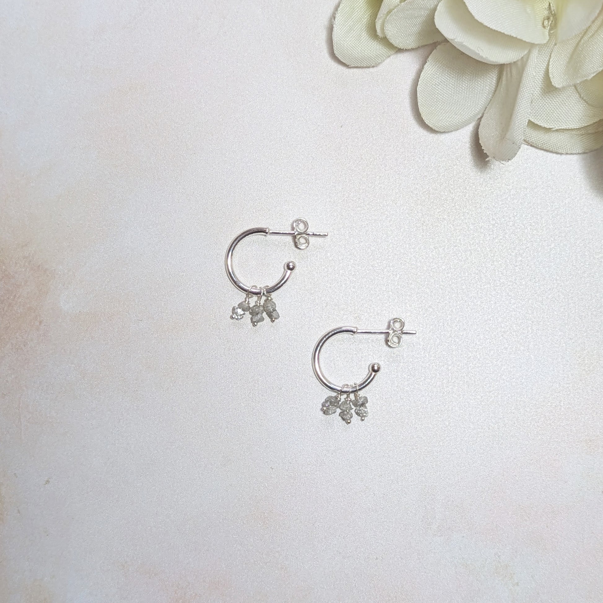 Silver hoop earrings with mini rough Diamond nugget charms on a white surface with a white flower.