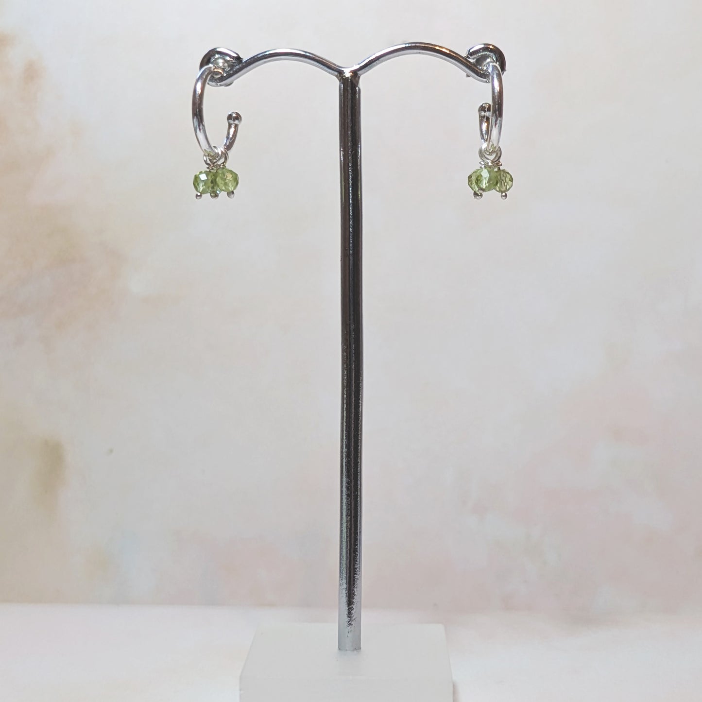 Pair of silver hoop earrings with green Peridot drops on a stand against a light background.