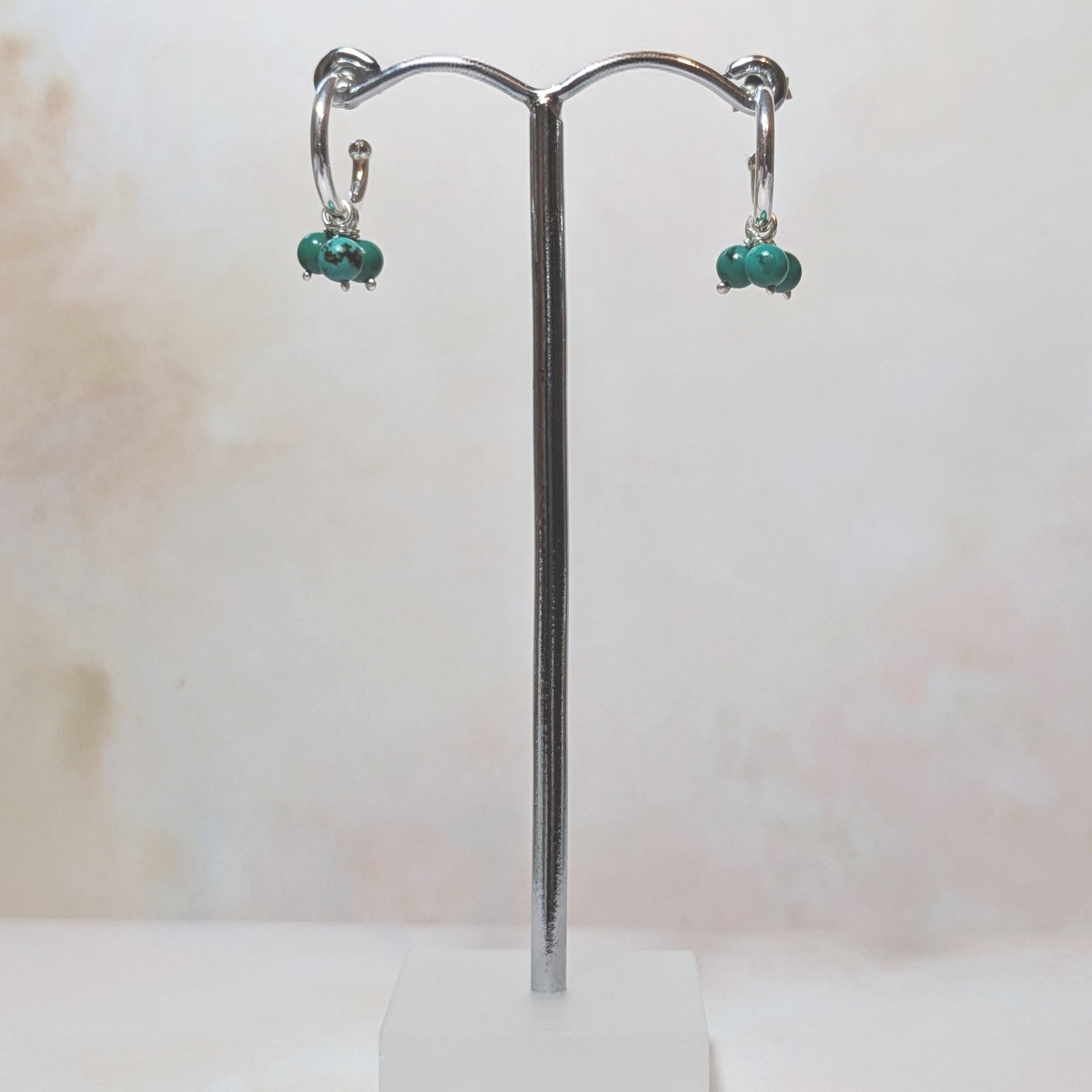 Pair of silver hoop earrings with natural Turquoise gemstone drop charms on a stand against a pale neutral background.