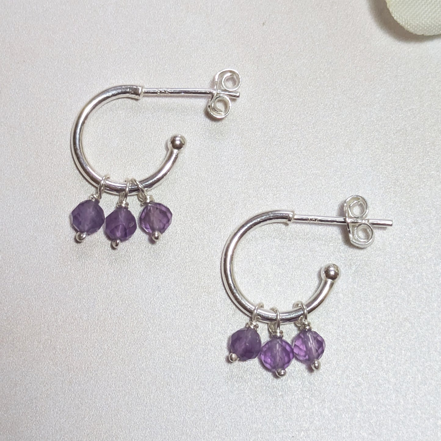 Silver hoop earrings with purple Amethyst gemstone charms on a light grey background.