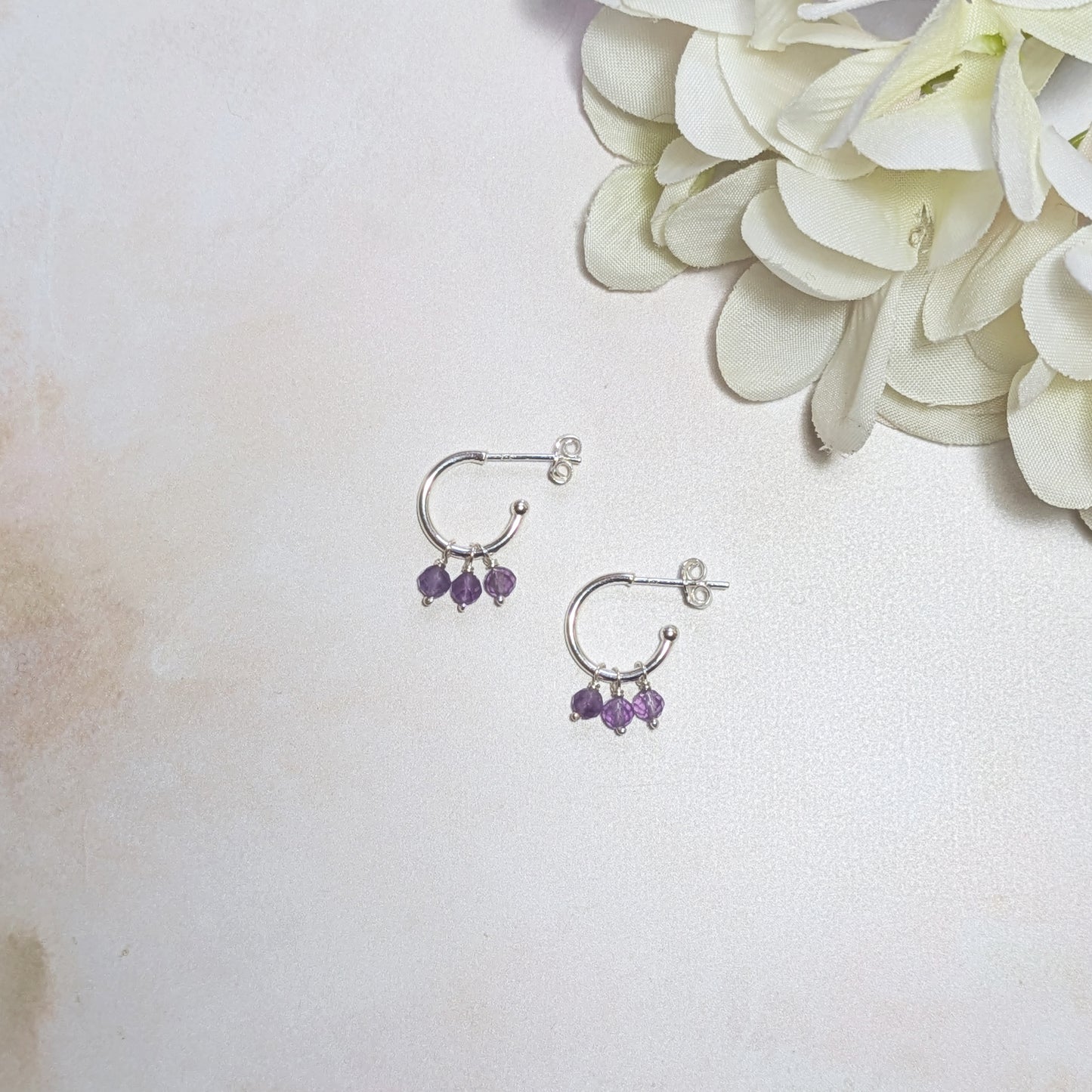 Sterling Silver hoop earrings with Amethyst beads on a light surface with white flowers.