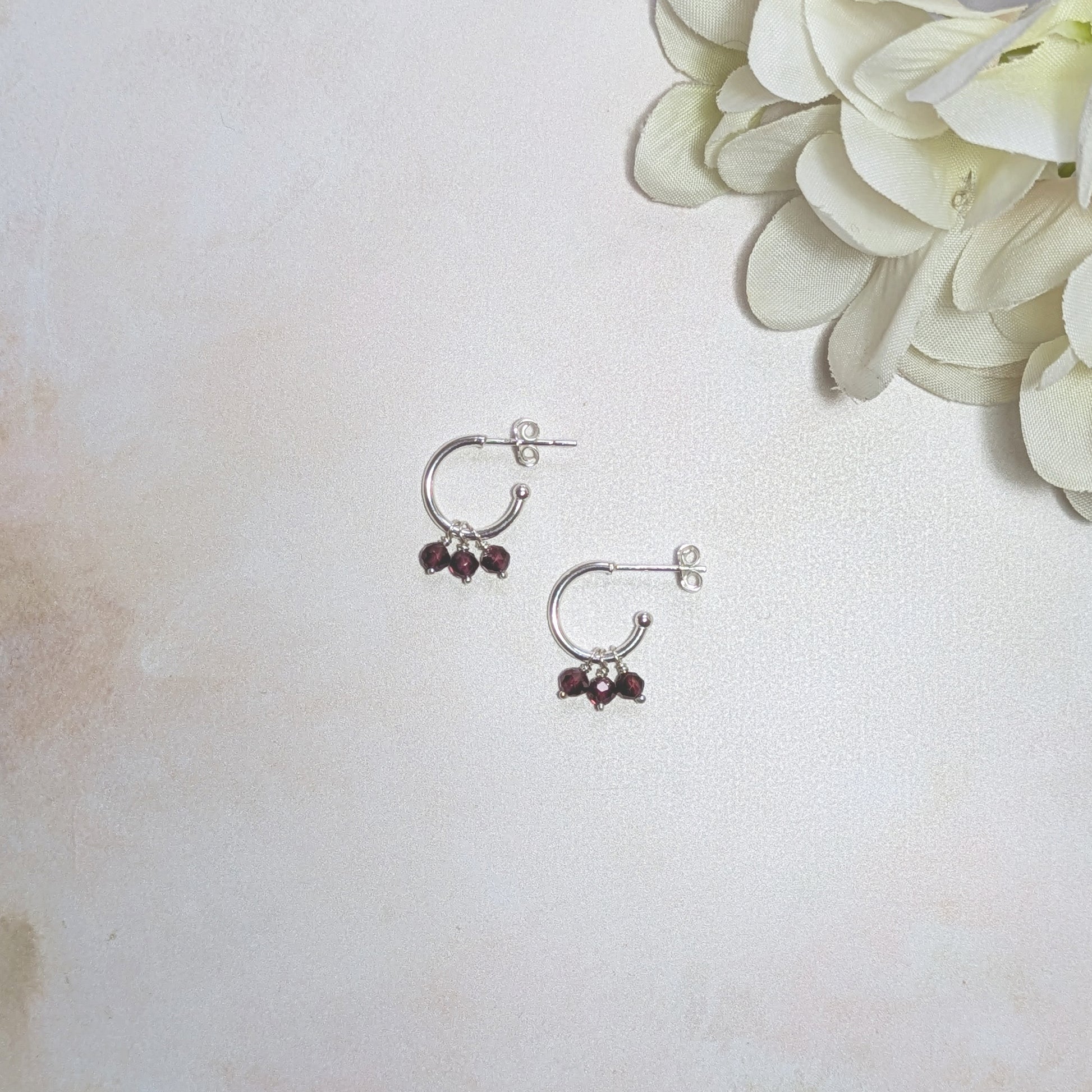 Silver hoop earrings with red Garnet gemstones on a light background with a white flower.