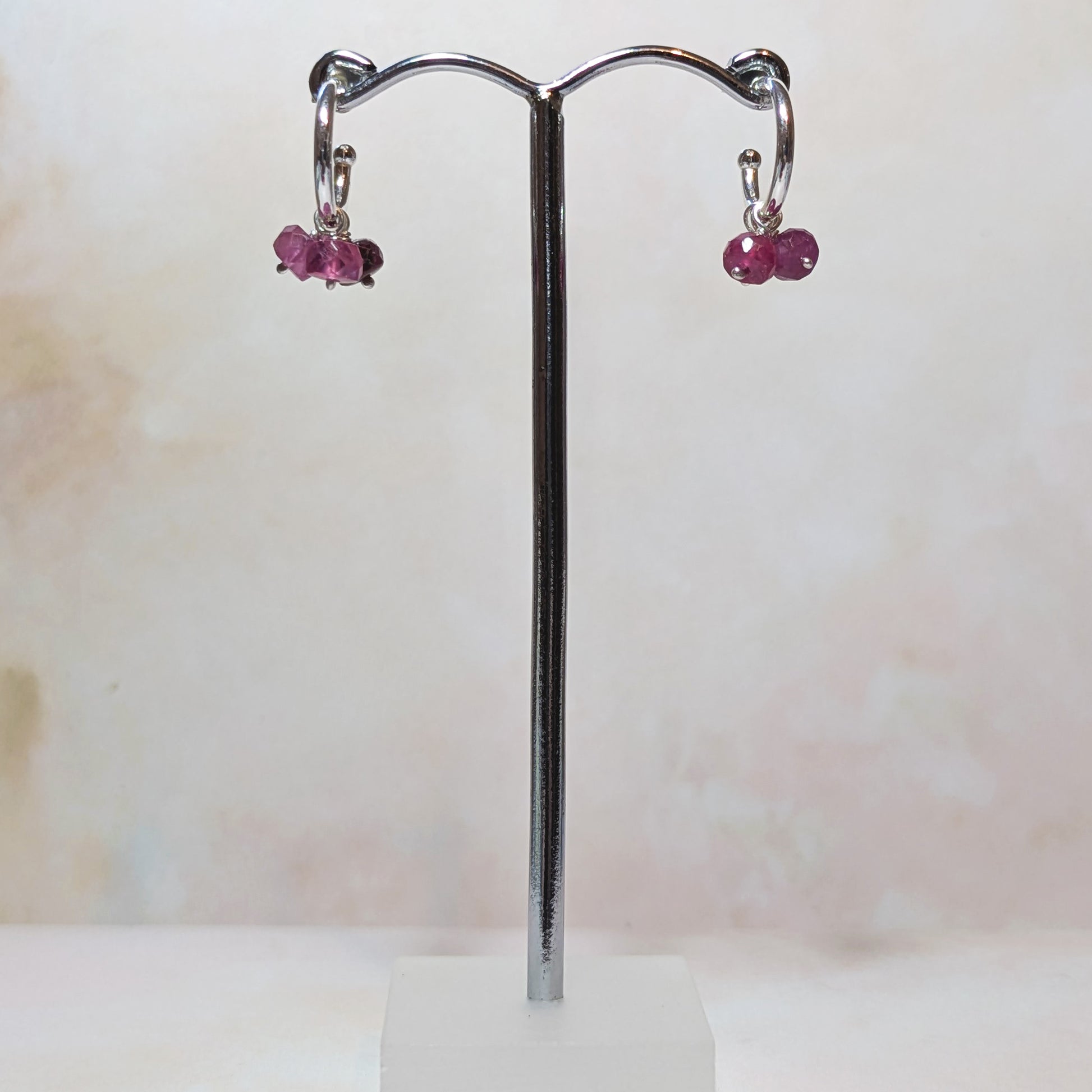 Pair of silver hoop earrings with Ruby gemstone charms on a stand against a light background.