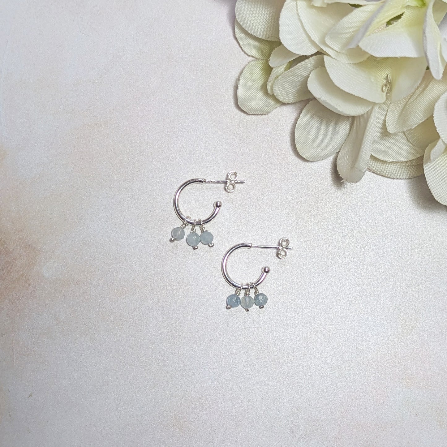 Silver hoop earrings with mini Aquamarine gemstones on a light background with a white flower.