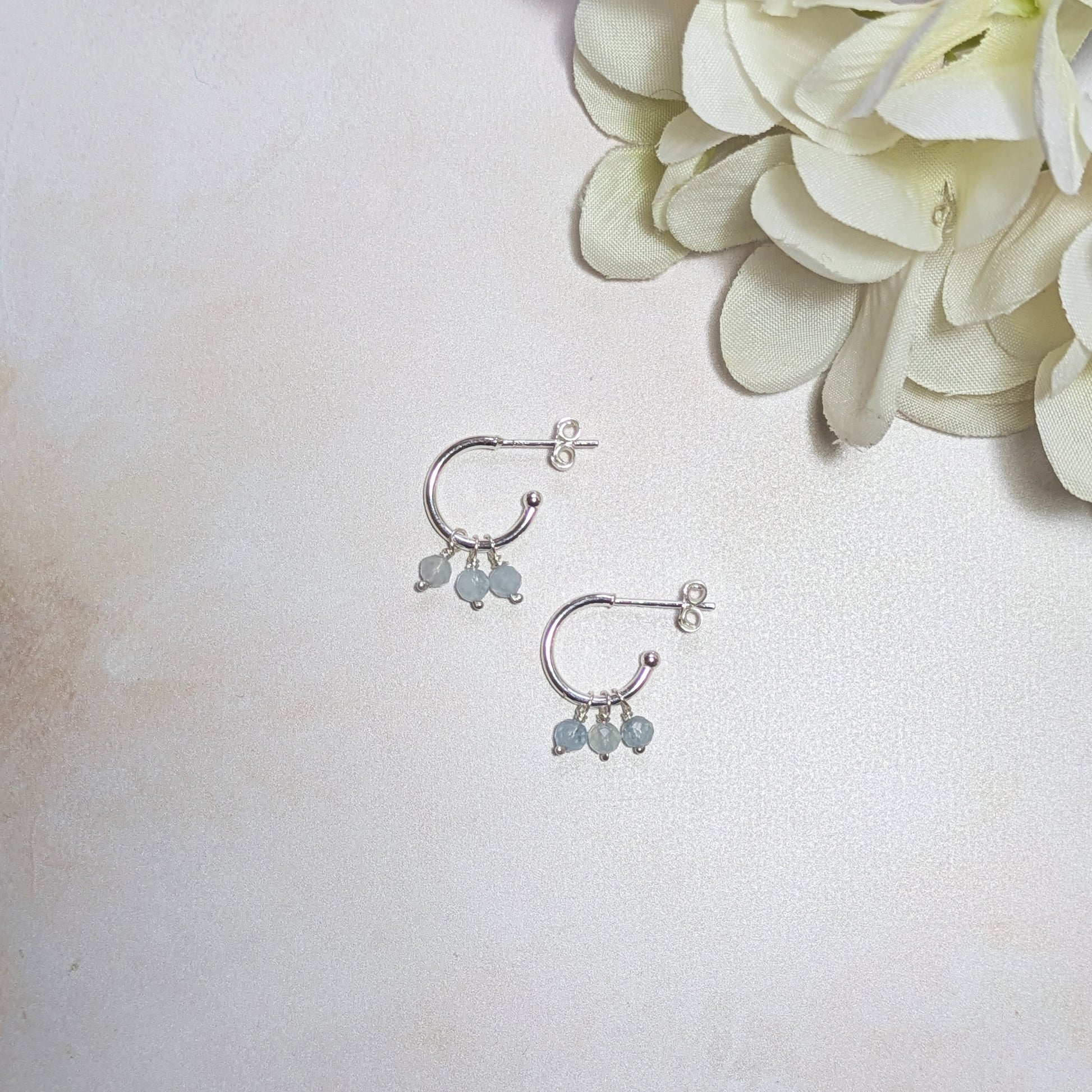 Silver hoop earrings with mini Aquamarine gemstones on a light background with a white flower.