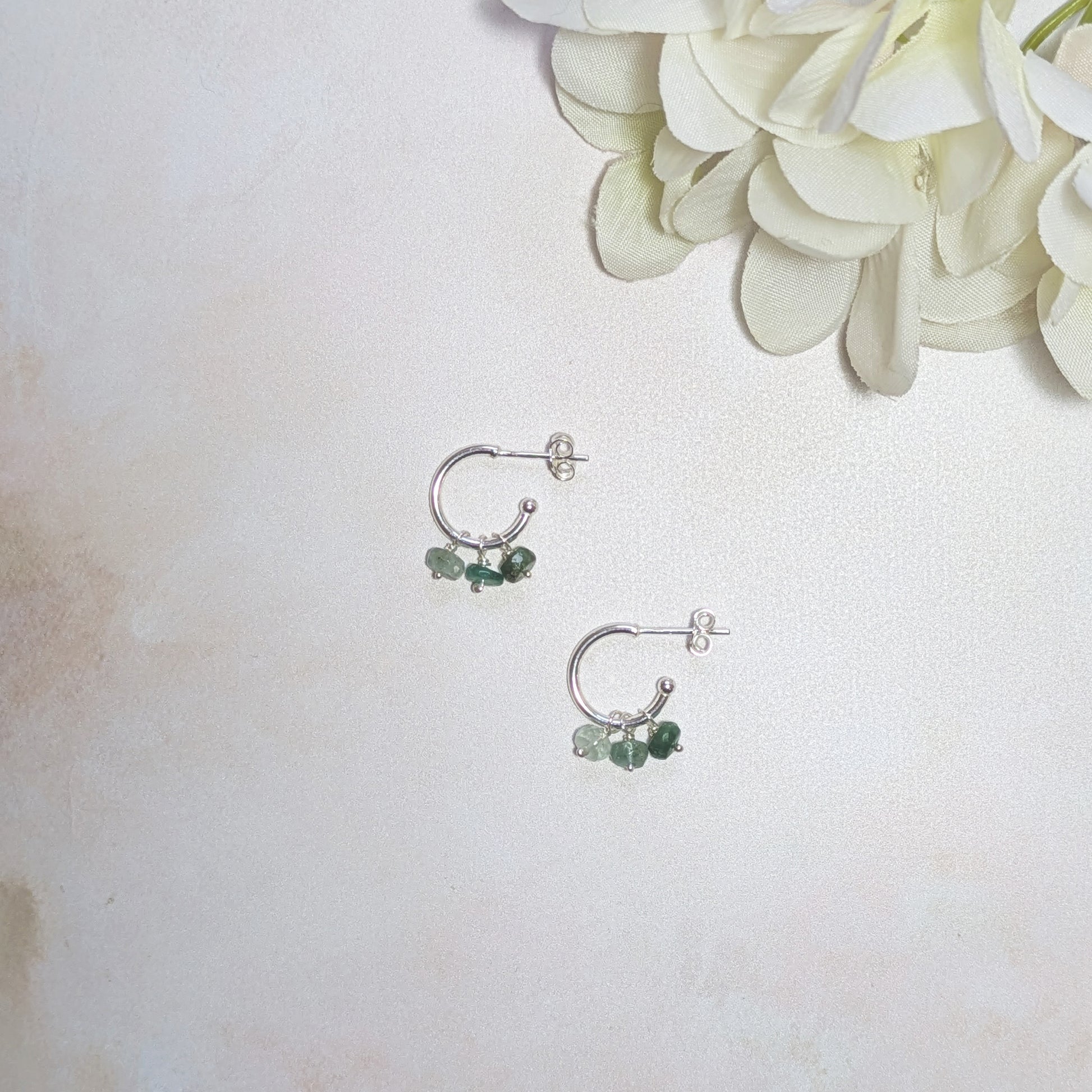 Pair of silver hoop earrings with Emerald gemstones on a light background with white flowers.