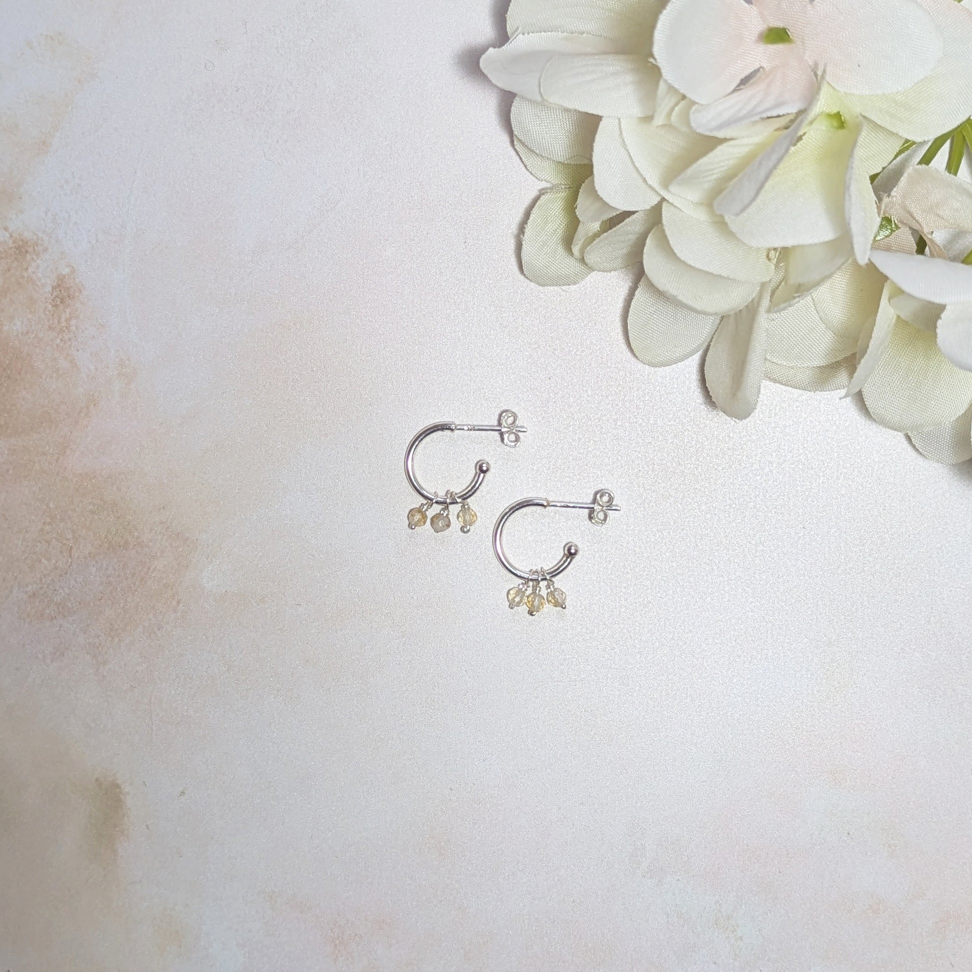 Silver hoop earrings with small dangling Citrine gemstone charms on a light background with white flowers.