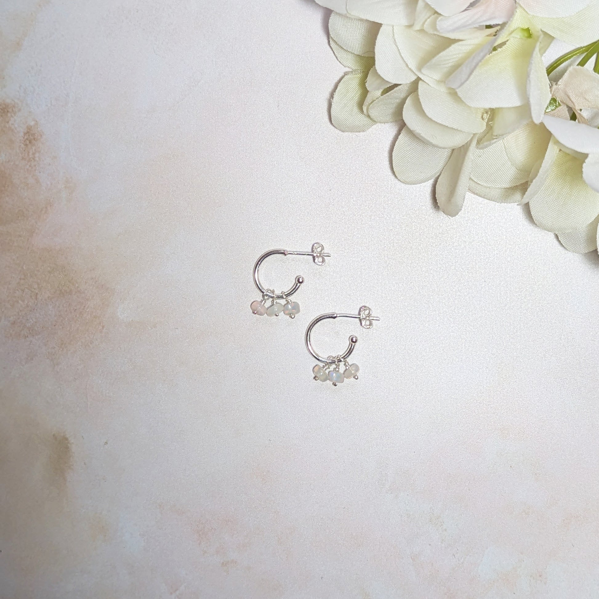Silver hoop earrings with small Opal gemstone drops on a pale neutral backround with white flowers.