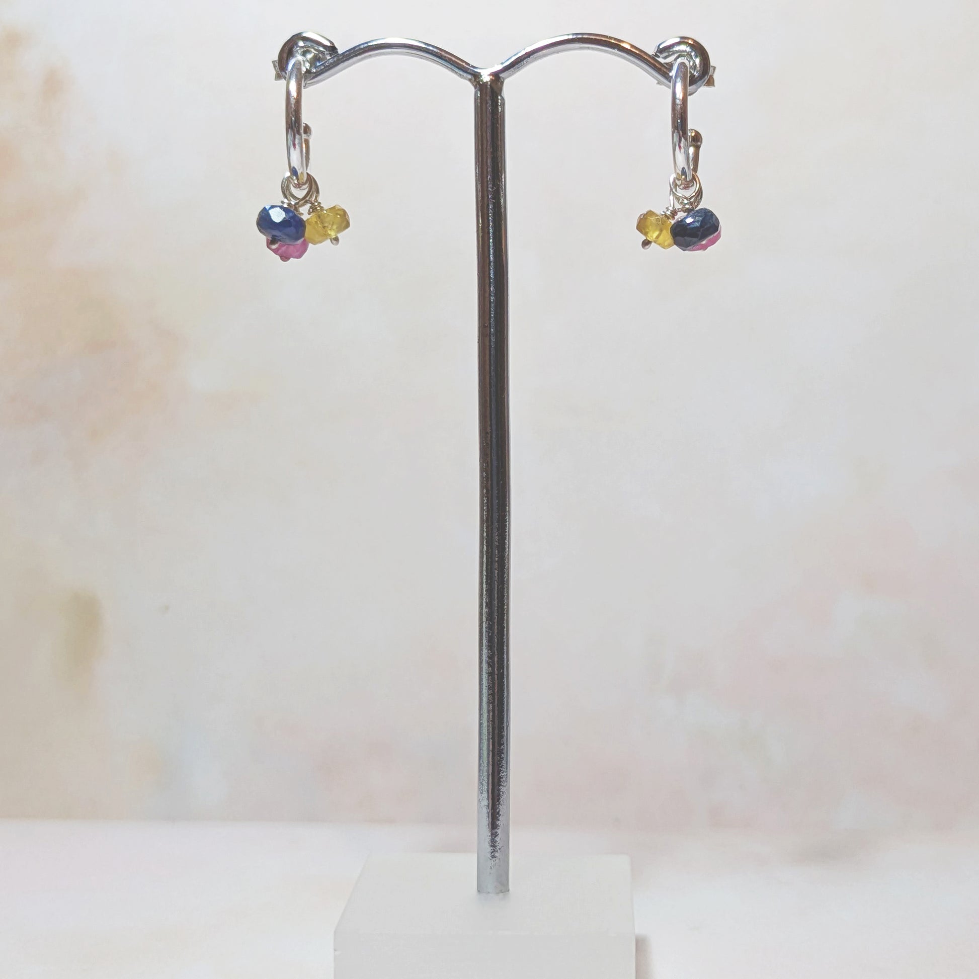 Pair of silver hoop earrings with multicoloured Sapphire drops on a stand against a light background.