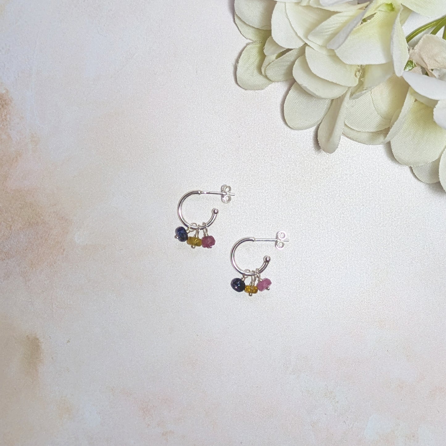 Pair of small hoop earrings with blue, pink and yellow Sapphire beads on a light background with flowers.