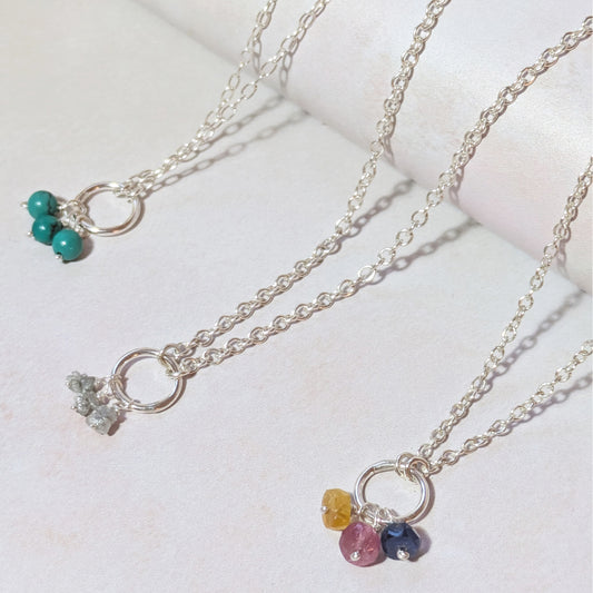 Three silver mini birthstone charm necklaces with gemstone pendants on a light background.