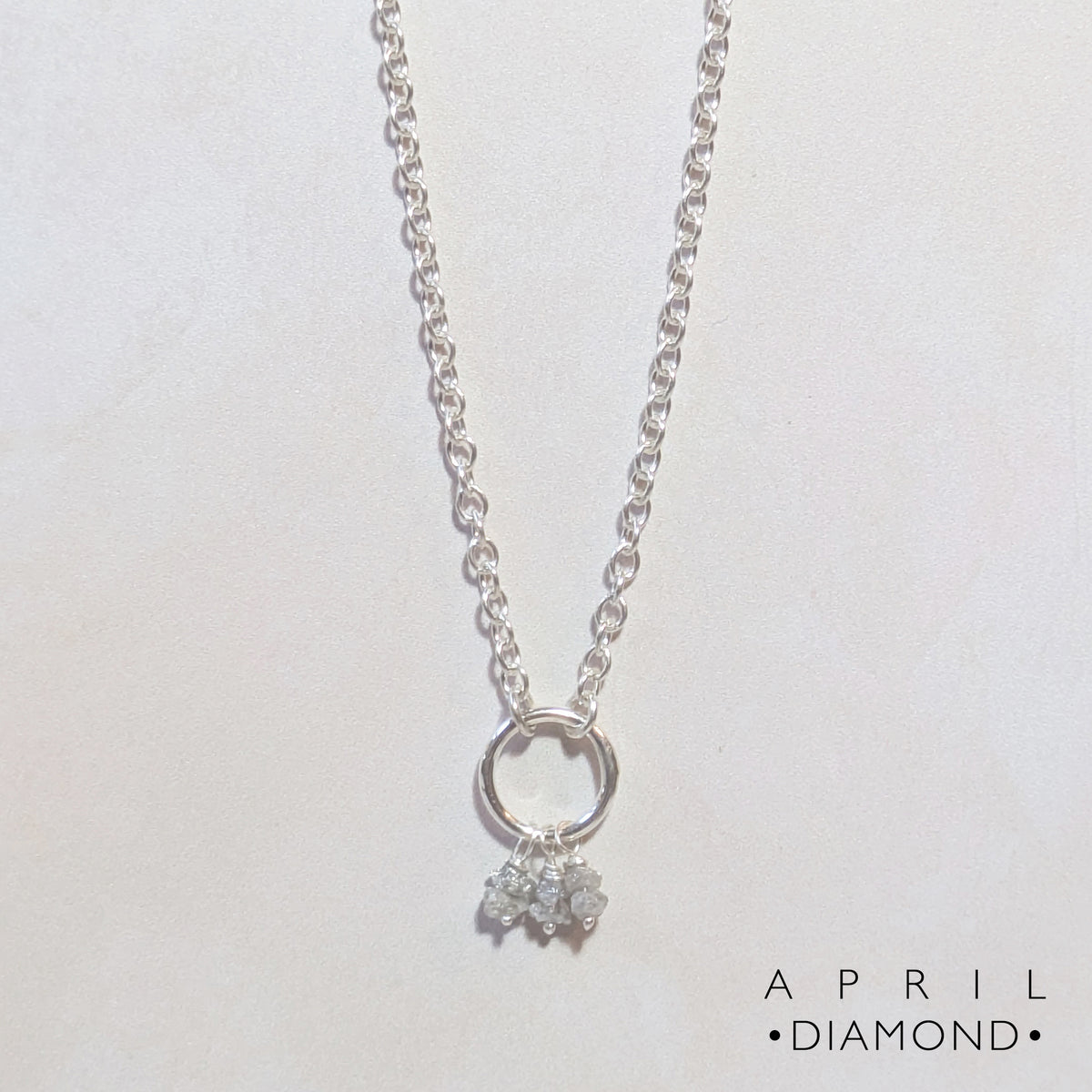 A silver chain birthstone necklace for April with a small silver circle pendant and a cluster Diamond nugget charms, shown on a plain light background. The text 'APRIL DIAMOND' is printed in the bottom right corner.