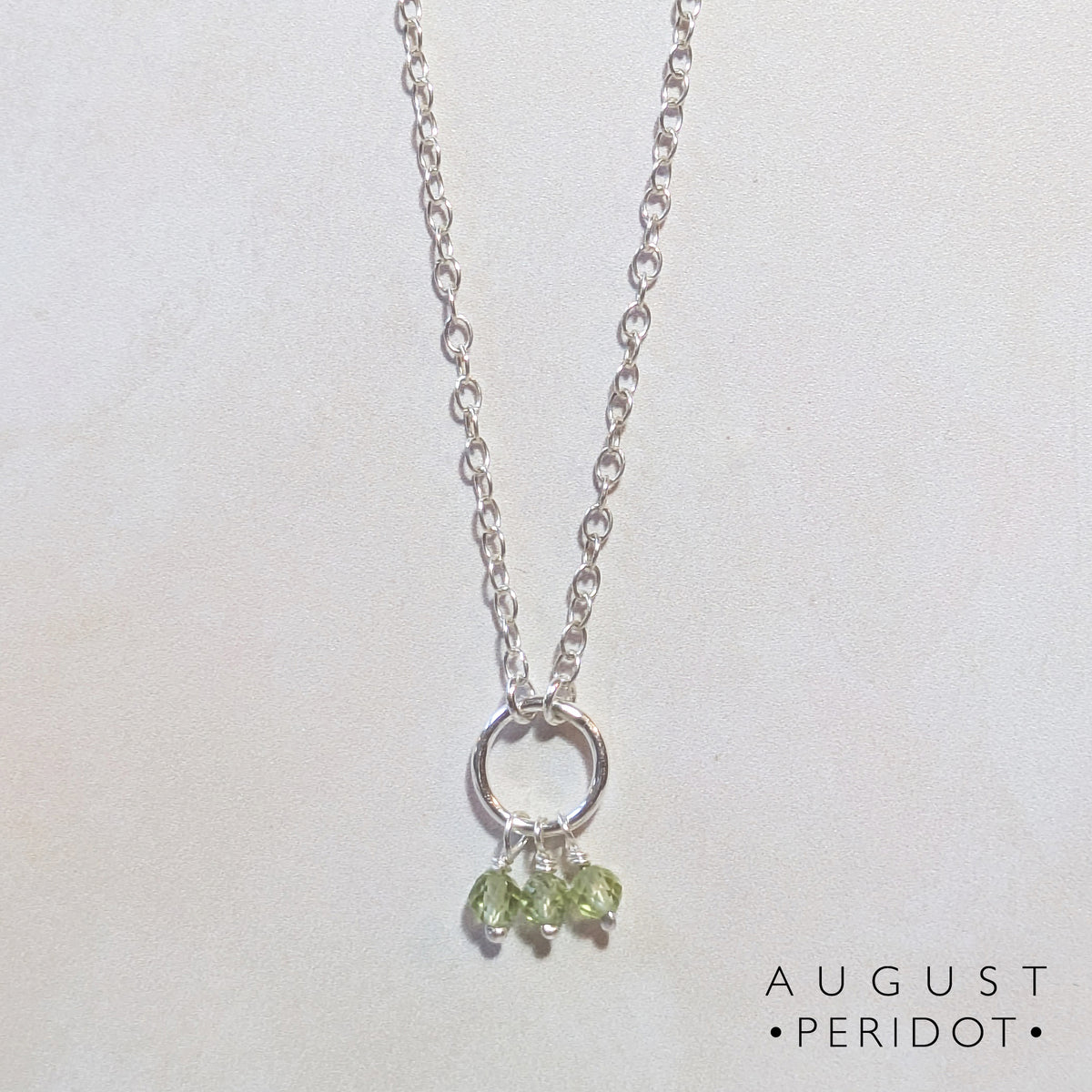 A silver chain birthstone necklace for August with a small silver circle pendant and three dangling light green peridot gemstones, shown on a plain light background with the text 'AUGUST PERIDOT' in the lower right.