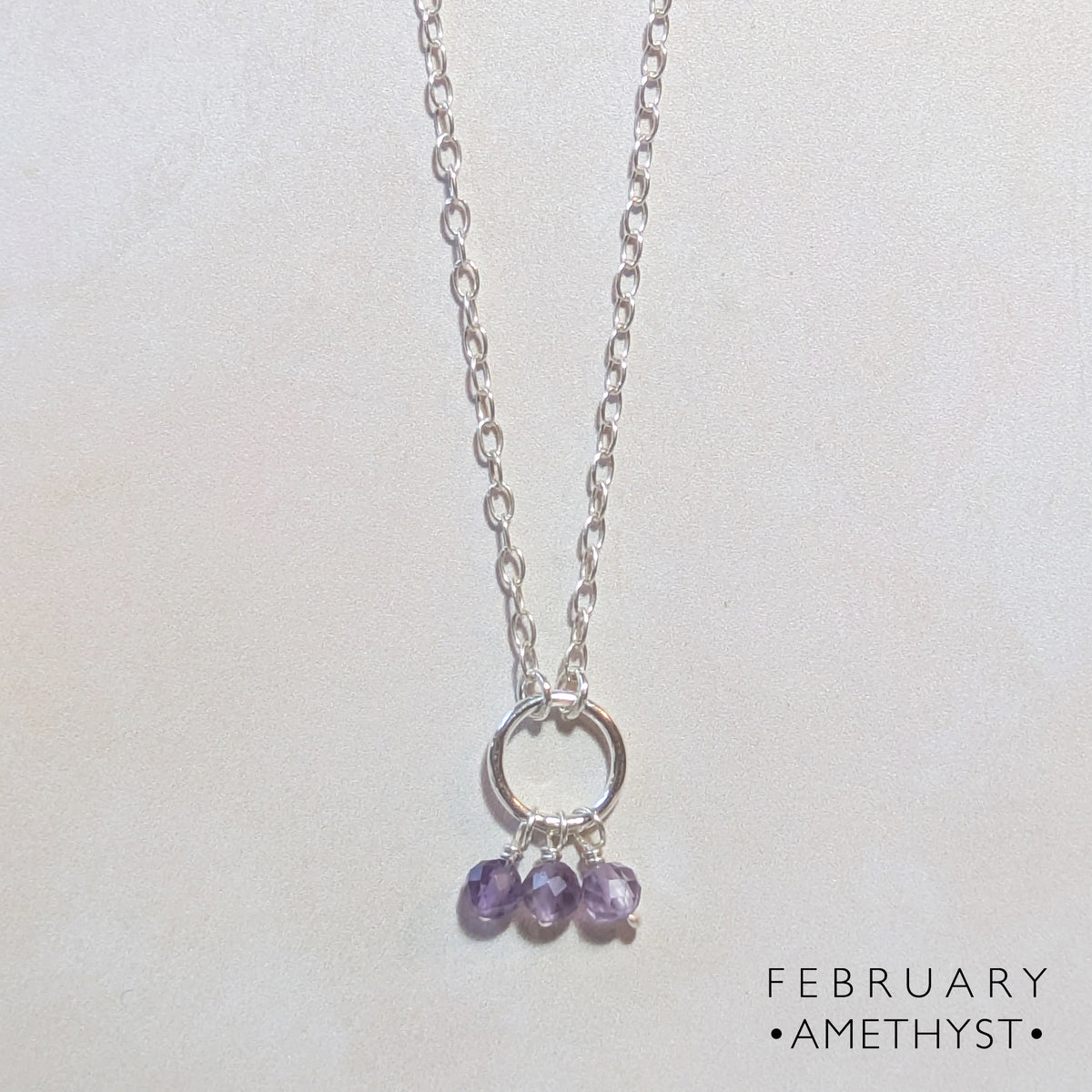 A silver chain birthstone necklace for February with a small circle pendant and three dangling amethyst gemstones, shown on a light background with the text 'FEBRUARY • AMETHYST •' in the lower right corner.