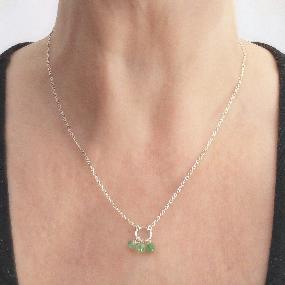 A close-up of a woman wearing a delicate silver chain birthstone necklace with a small silver circle and three small Emerald bead charms.
