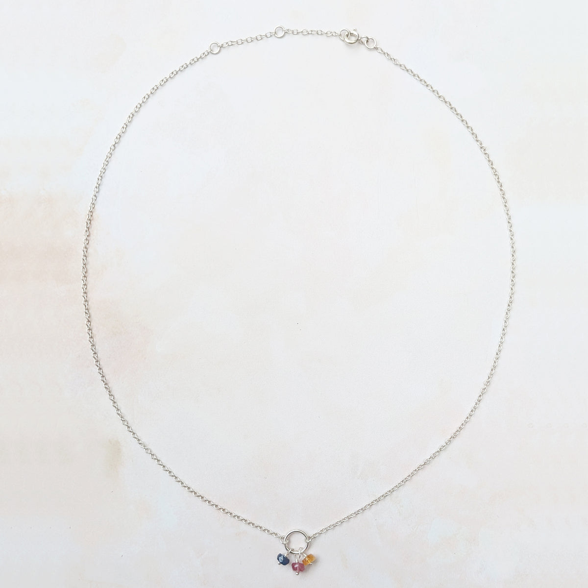 A delicate silver chain necklace with a small circle and three multicoloured Sapphire gemstone charms in blue, pink, and yellow, displayed on a light cream marble background.