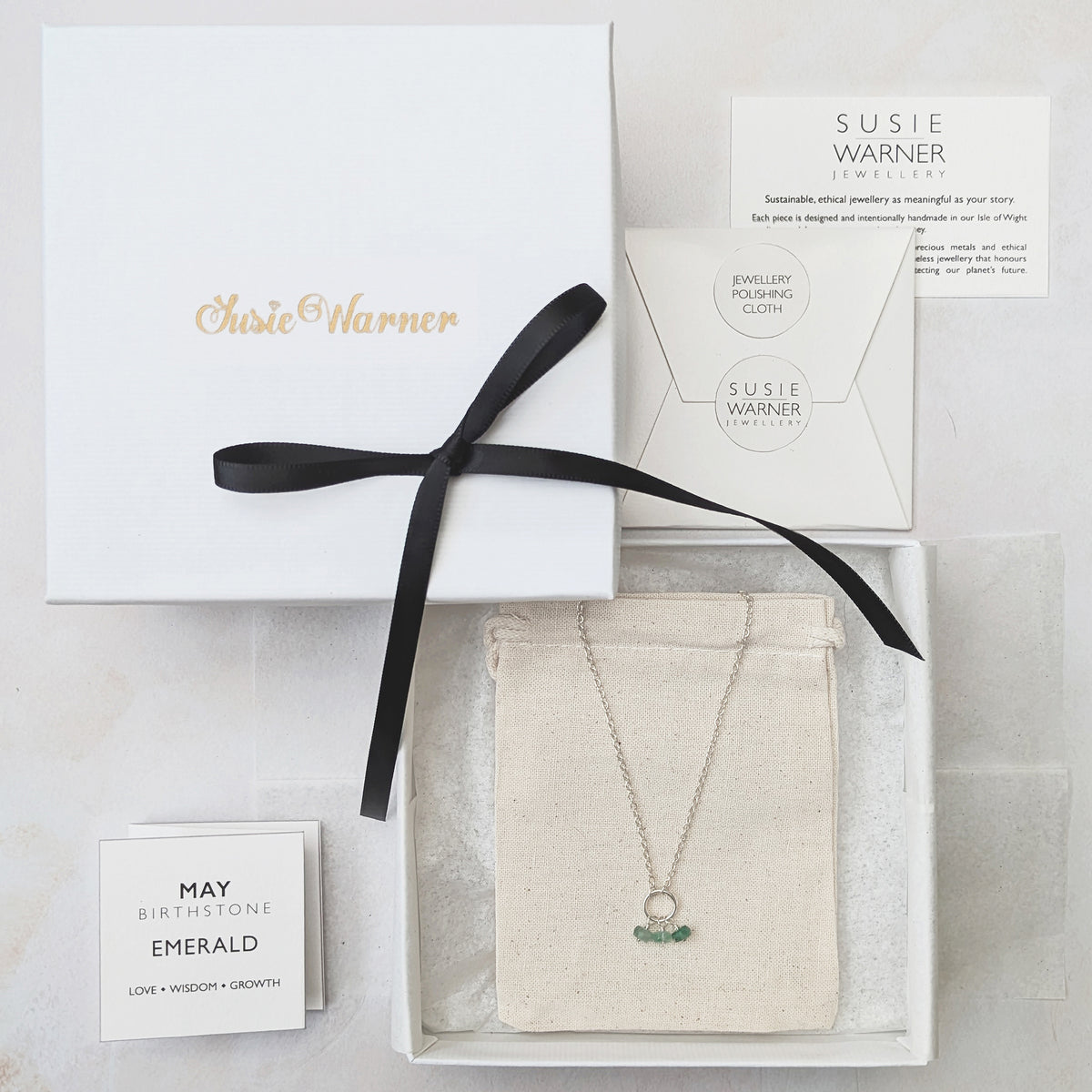 A gift set from Susie Warner Jewellery featuring a silver necklace with a green emerald pendant, presented in a white box with a black ribbon, alongside a white card and a branded box lid.
