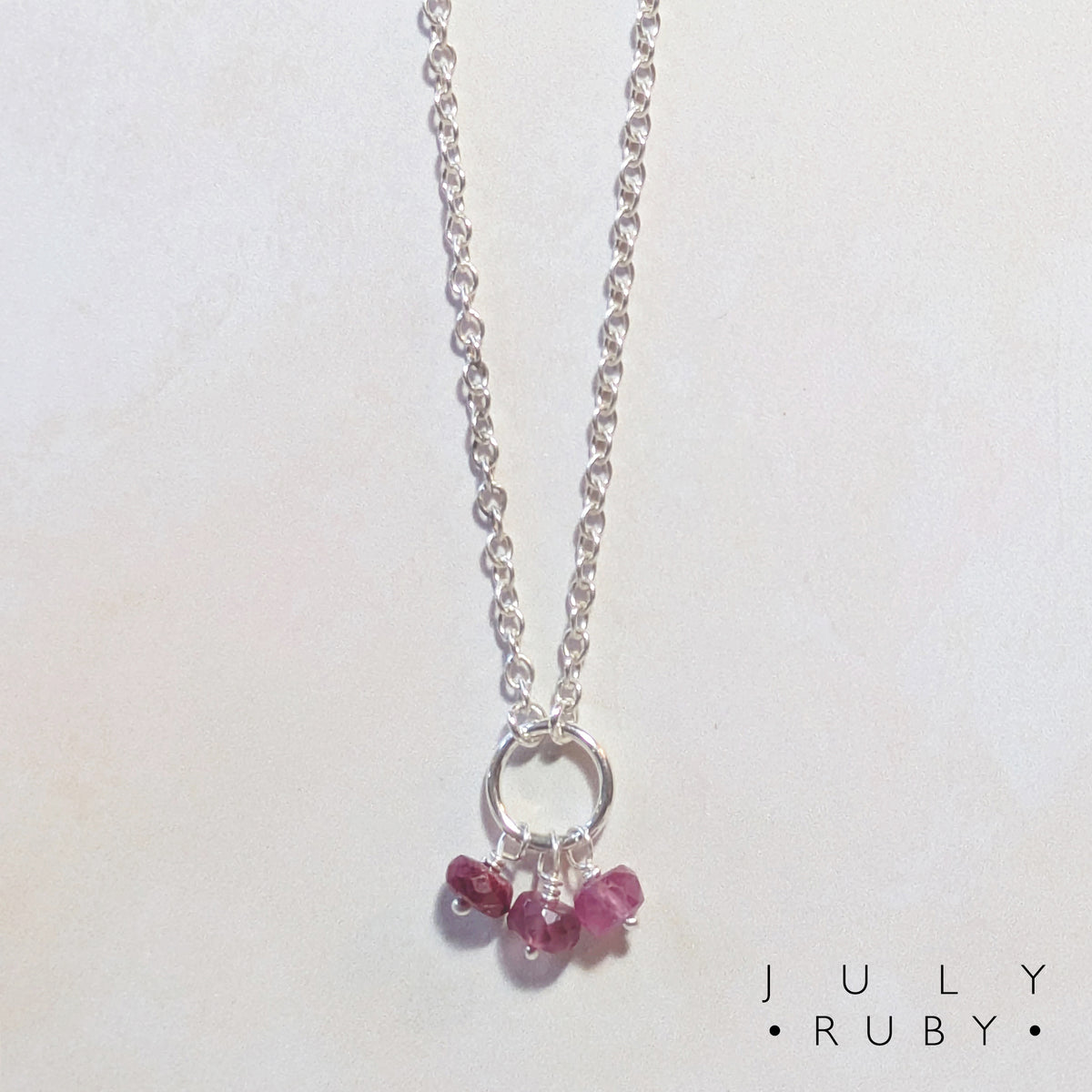 A silver chain birthstone necklace for July  with a small silver circle and three dangling pink Ruby gemstone charms, photographed on a plain light background. The text 'JULY RUBY' appears in the lower right corner.