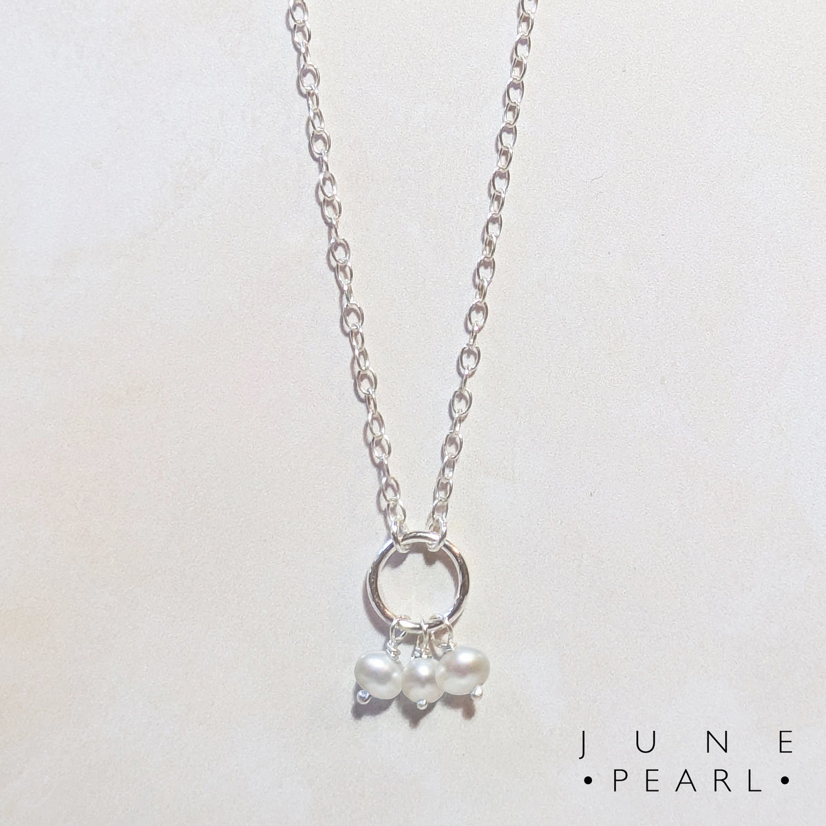 A silver chain birthstone necklace for June with a small circle pendant and three dangling white pearls, shown on a light beige background with the text 'JUNE PEARL' in the bottom right corner.
