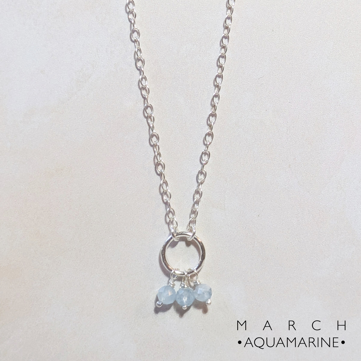 A silver chain birthstone necklace for March with a small circle pendant and three dangling light blue aquamarine gemstones, displayed on a light beige background with 'MARCH AQUAMARINE' text in the lower right.
