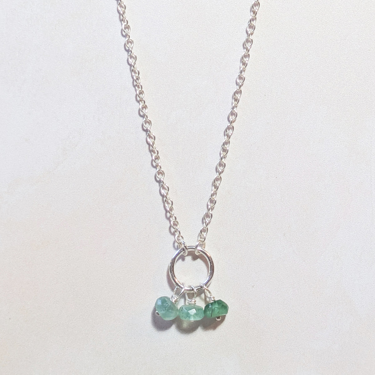 A delicate silver chain May birthstone necklace featuring a small silver circle pendant with three small, faceted natural Emerald gemstones hanging from it, displayed on a plain off-white background.
