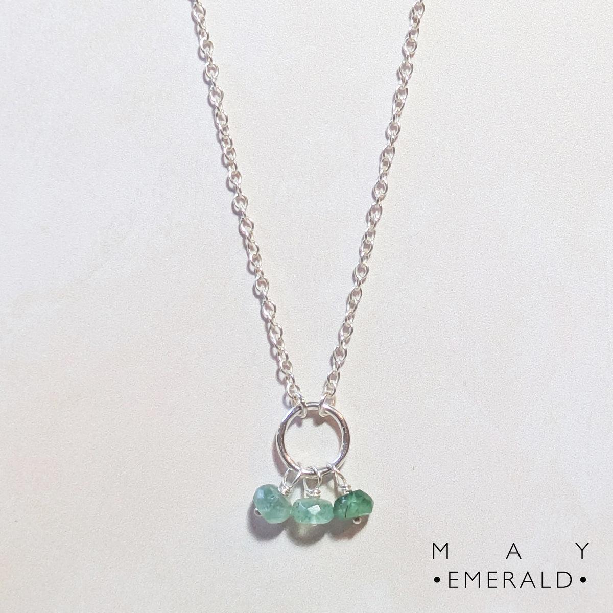 A silver chain birthstone necklace for May with a circle pendant holding three small Emerald faceted gemstones, displayed on a light off-white background. The text 'MAY EMERALD' is printed in the bottom right corner.