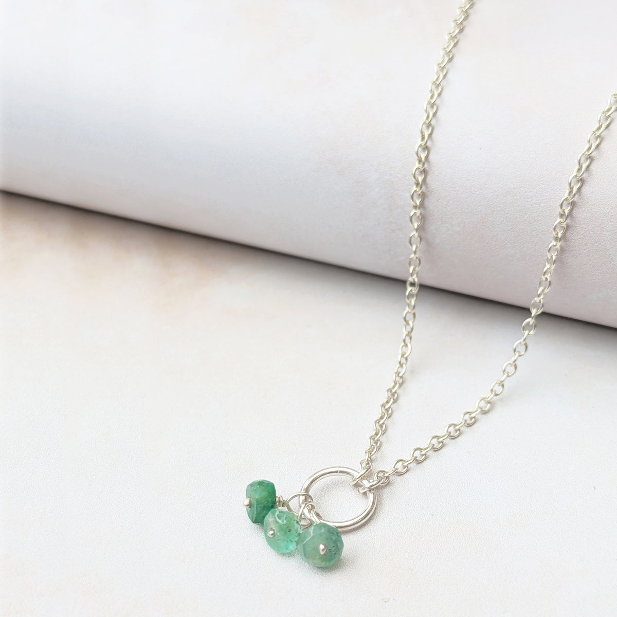 A delicate silver chain necklace featuring a small silver circle and three dangling Emerald faceted beads, displayed on a soft white fabric surface.