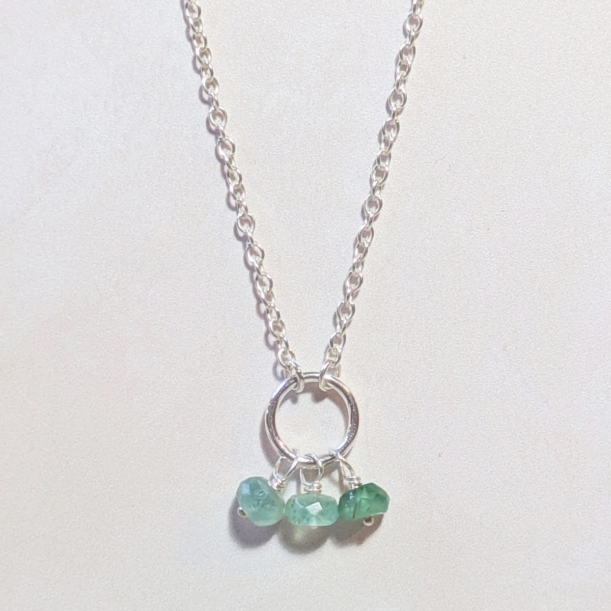A silver chain necklace with a circular ring pendant and three small Emerald faceted gemstones hanging from the bottom, displayed on a light off-white background.
