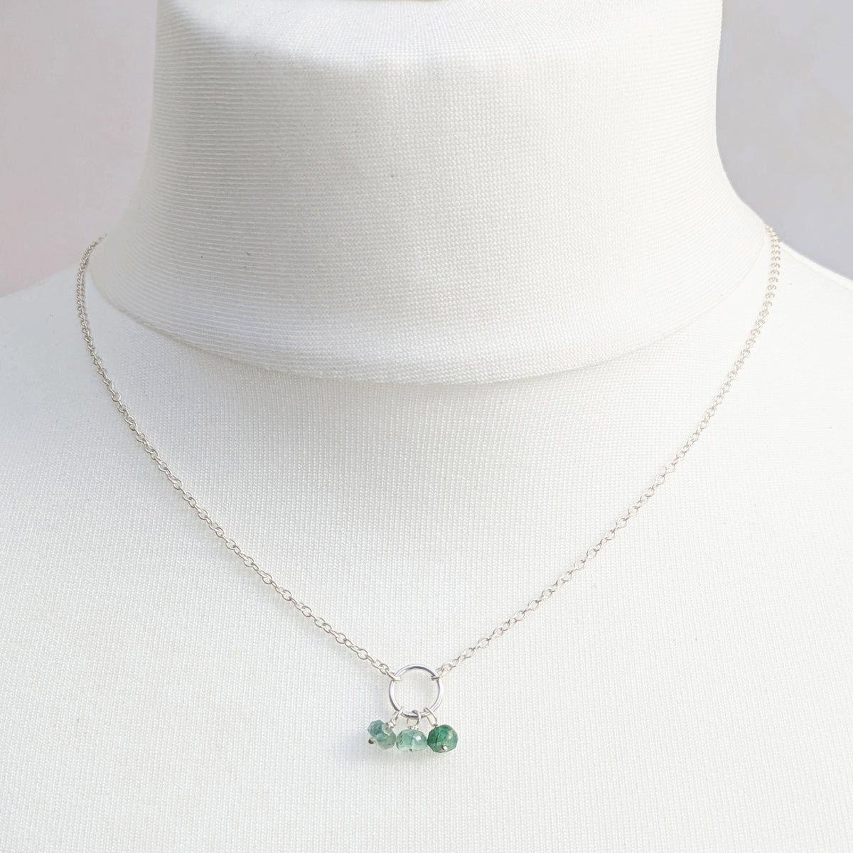 A delicate silver chain necklace with a small silver circle and three small Emerald faceted beads, displayed on a white fabric mannequin bust.