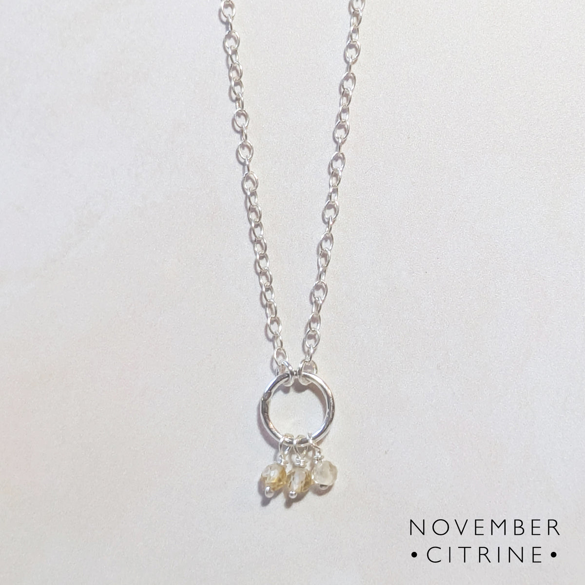 A silver chain birthstone necklace for November with a circle pendant and three small dangling citrine gemstones, displayed on a light off-white background with the text 'NOVEMBER CITRINE' in the bottom right corner.