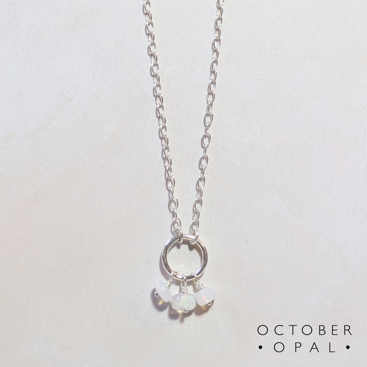 A silver chain birthstone necklace  for October with a circle pendant and three small natural opal beads, shown on a plain light background. The text 'OCTOBER OPAL' appears in the lower right corner.