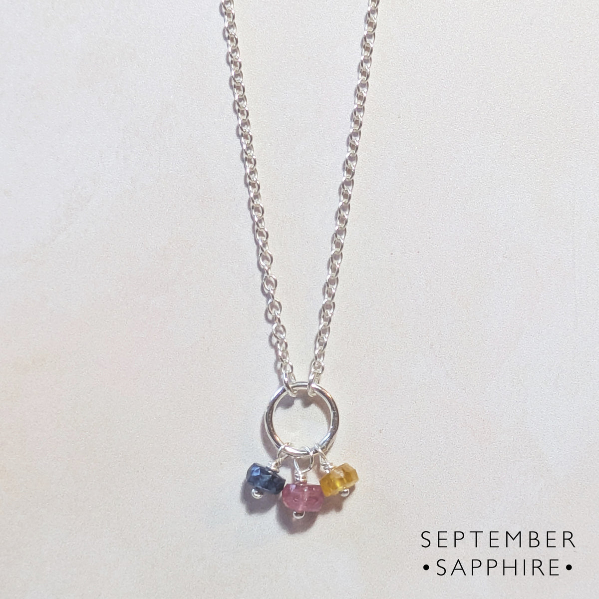 A silver chain birthstone necklace for September with a small circle pendant and three dangling multicoloured Sapphire gemstone charms in blue, pink, and yellow, shown on a plain light background with the text 'SEPTEMBER SAPPHIRE' in the lower right.